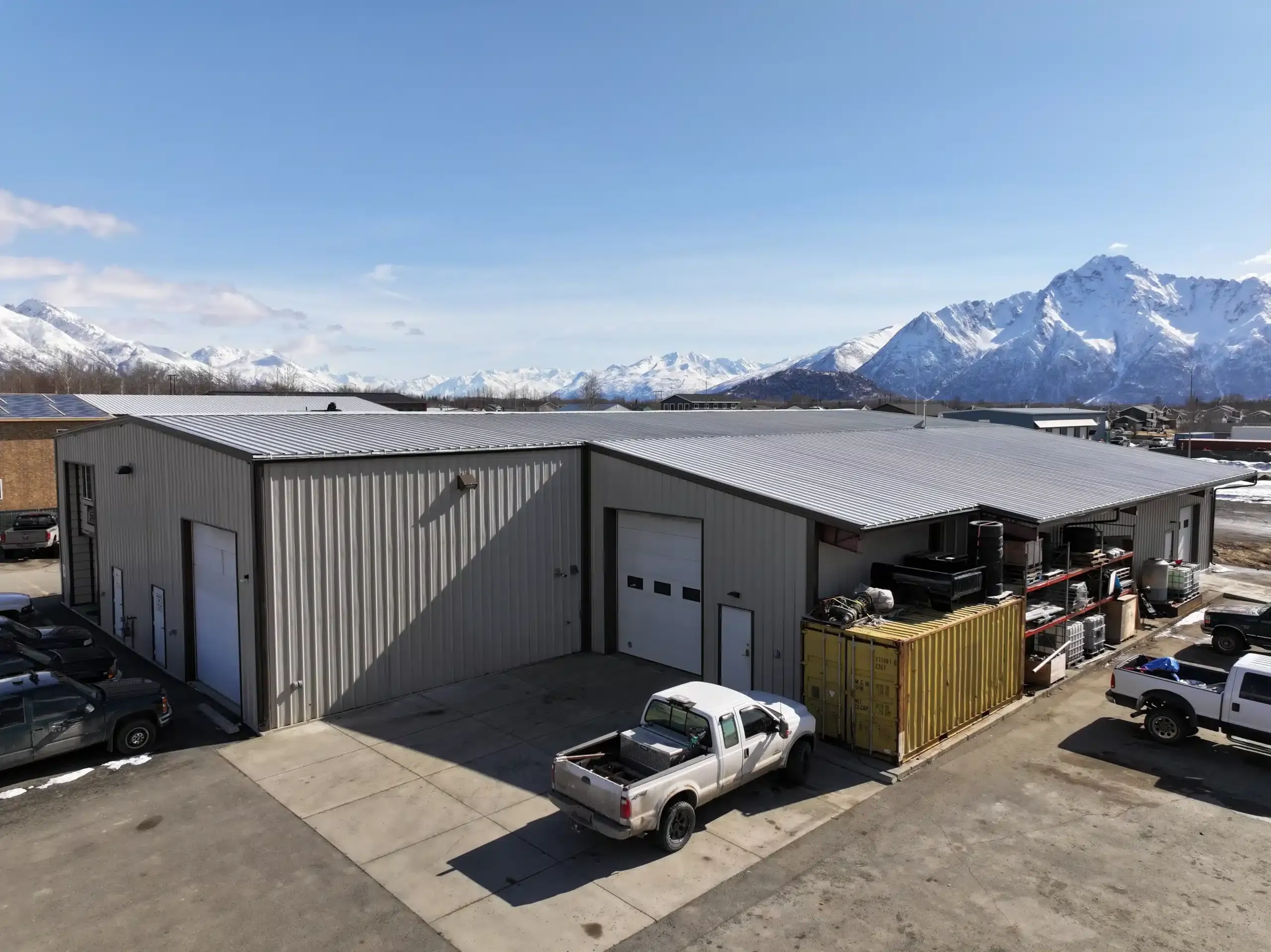 Industrial warehouse with a metal roof, surrounded by mountains. Several vehicles, including a white pickup truck, are parked outside. Clear blue sky and scattered snow patches complete the scene.