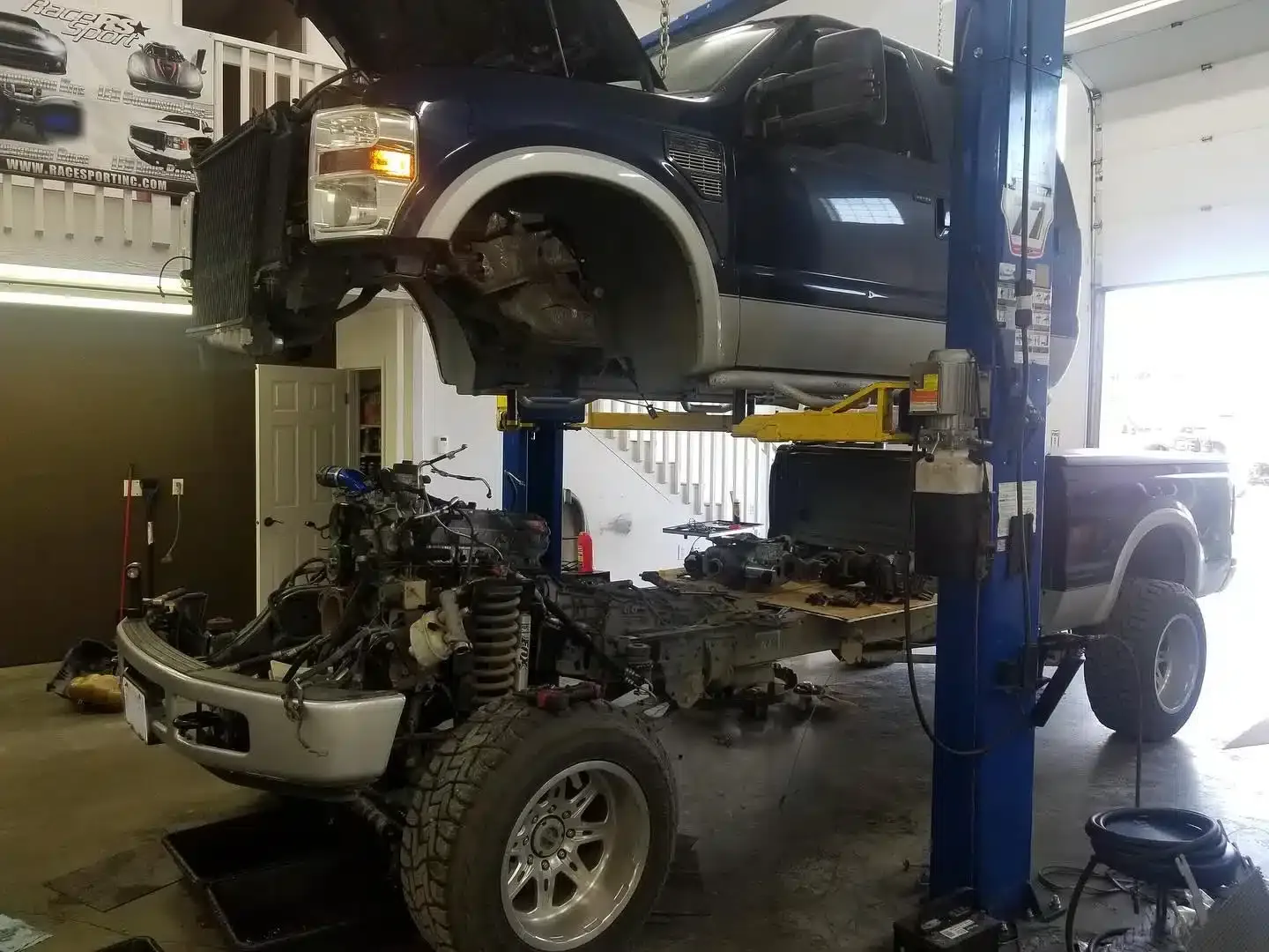 A blue truck is lifted on a hydraulic car lift in a garage, with its body raised and separated from the chassis. Exposed mechanical components are visible, and workshop tools are around the area.