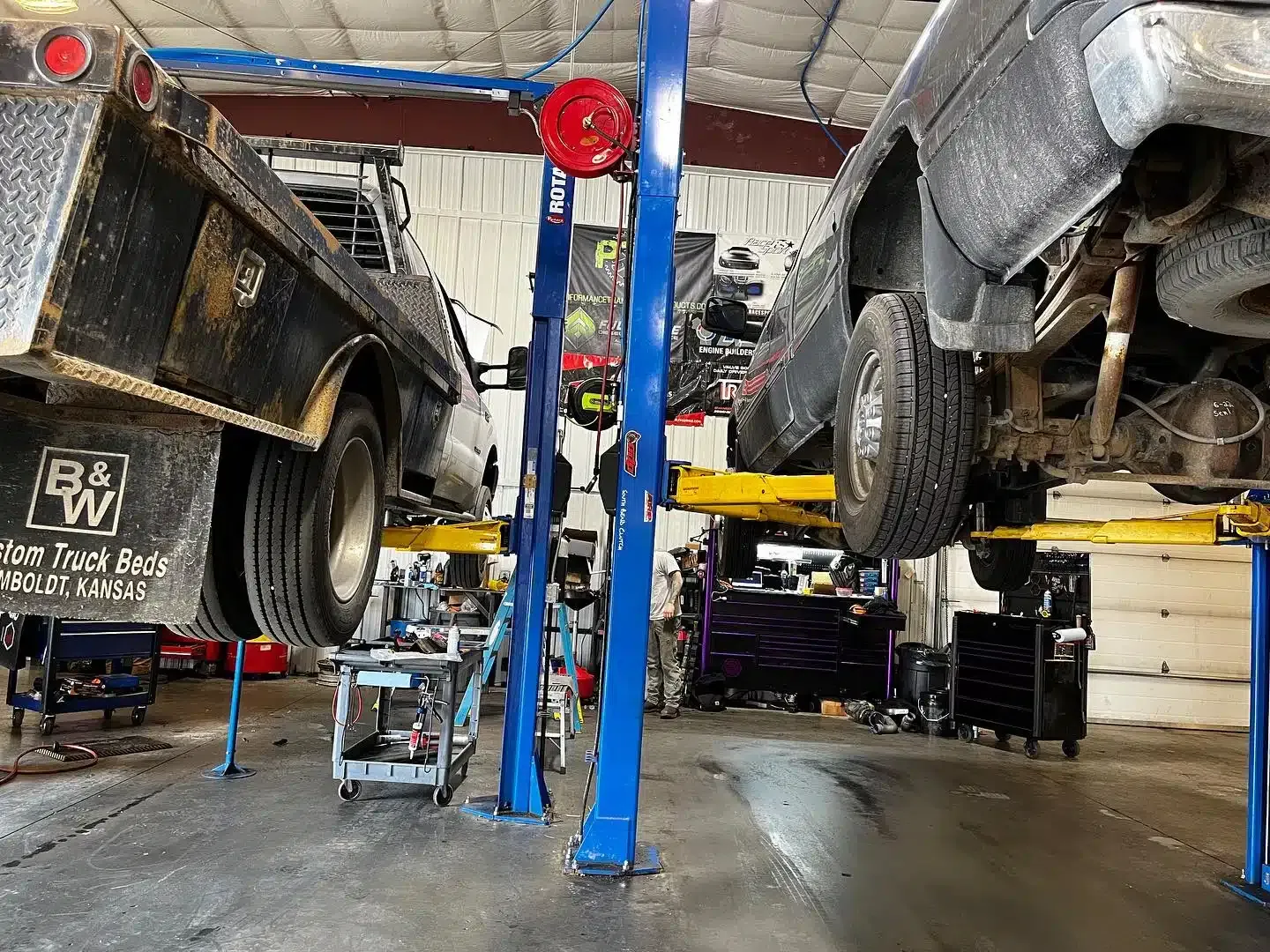Two trucks are elevated on hydraulic lifts inside a well-lit mechanic shop. A worker in the background is focused on repairing one truck. Various tools and equipment are organized throughout the workspace.