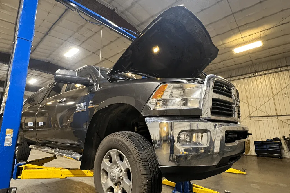 Diesel Maintenance in Palmer, AK at 1023 Diesel & Fleet. A large black pickup truck is lifted on a hydraulic car lift inside an automotive repair shop. The truck’s hood is open, and the garage is brightly lit with tools and equipment visible in the background
