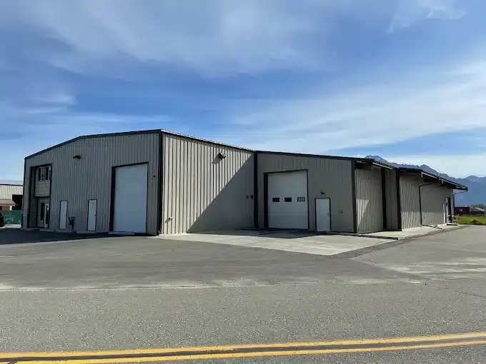 A large, empty industrial warehouse with gray metal siding and multiple closed roll-up doors. The building is situated on a paved lot under a clear blue sky. Mountains are faintly visible in the background.