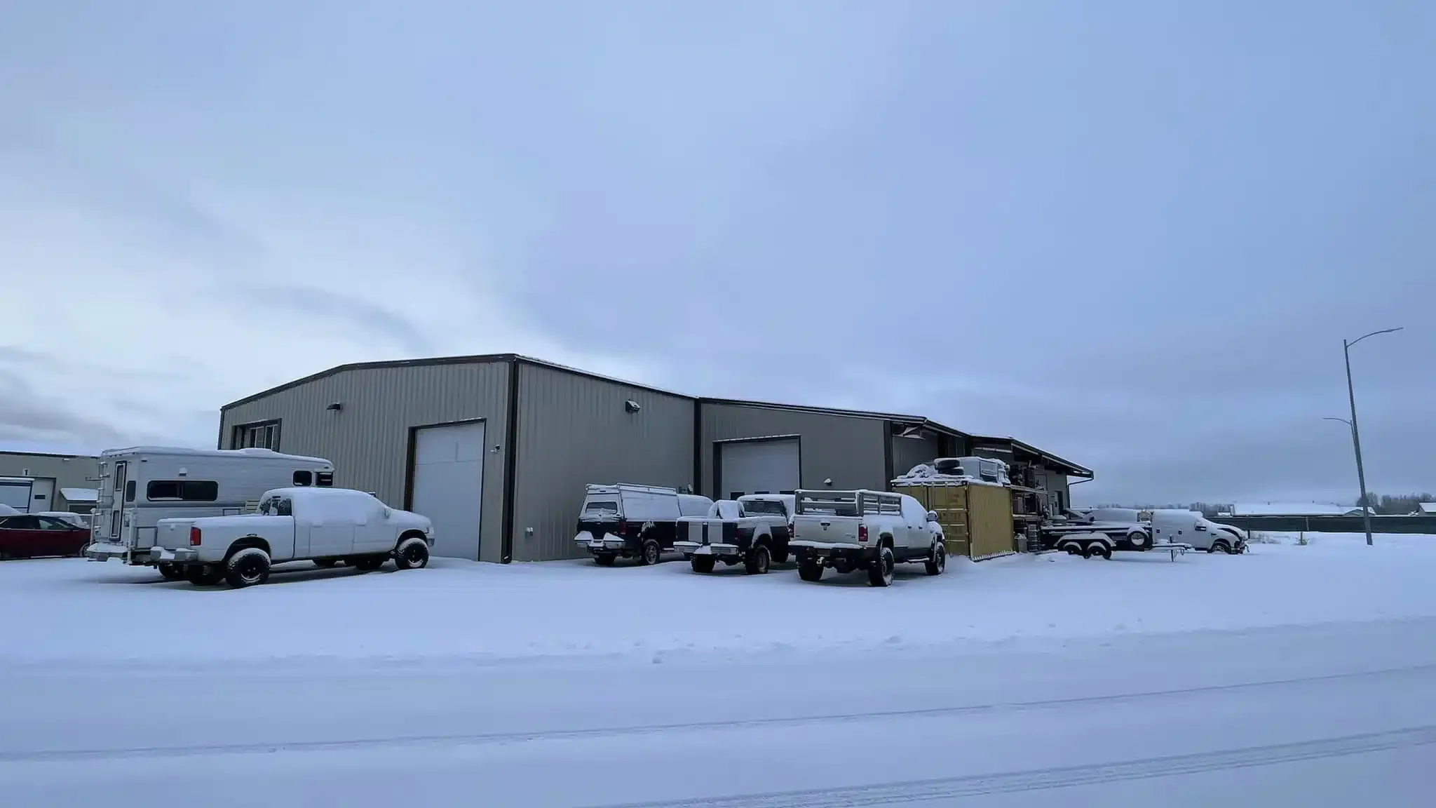 A snowy industrial area with several parked vehicles, including vans and trucks, beside a large, grey warehouse. The ground and rooftops are covered in snow, and the sky is overcast.