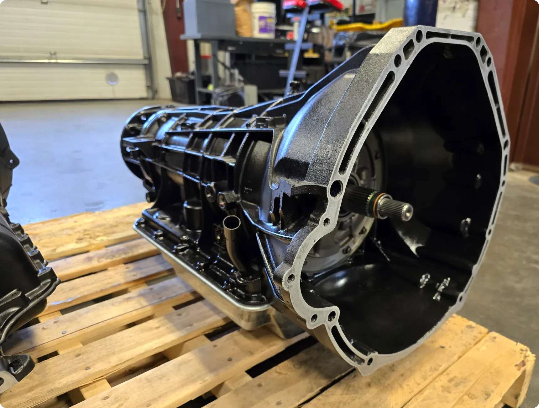 Transmissions rebuild service 1023 Diesel & Fleet: Close-up of a black, heavy-duty vehicle transmission on a wooden pallet in a workshop setting, with tools and equipment visible in the background.