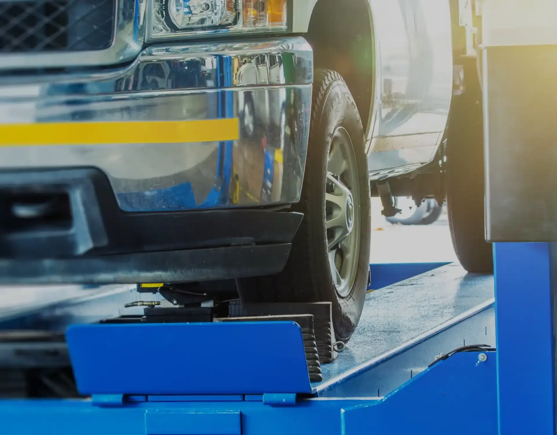 1023 Diesel & Fleet: A vehicle is positioned on a blue hydraulic lift inside an auto repair shop. Sunlight illuminates the scene, highlighting the front wheel and bumper of the car.