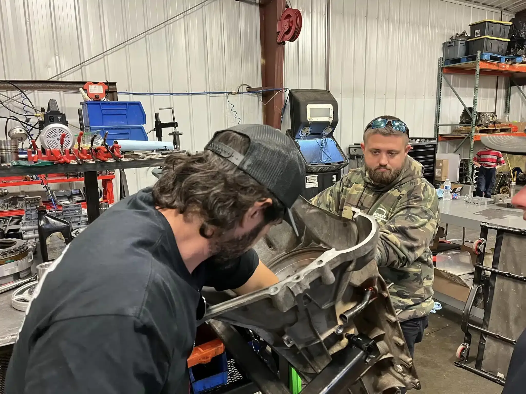 Two men are working together in a garage. One is focused on repairing an engine part on a workbench, while the other watches closely. Various tools and equipment are visible in the background.