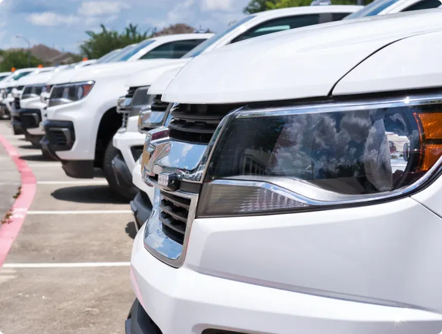 1023 Diesel & Fleet: A row of white pickup trucks parked in a lot, viewed from the side with a focus on the front headlights and grilles. The sky is partly cloudy and reflects on the truck surfaces. Trees and parked vehicles are visible in the background.