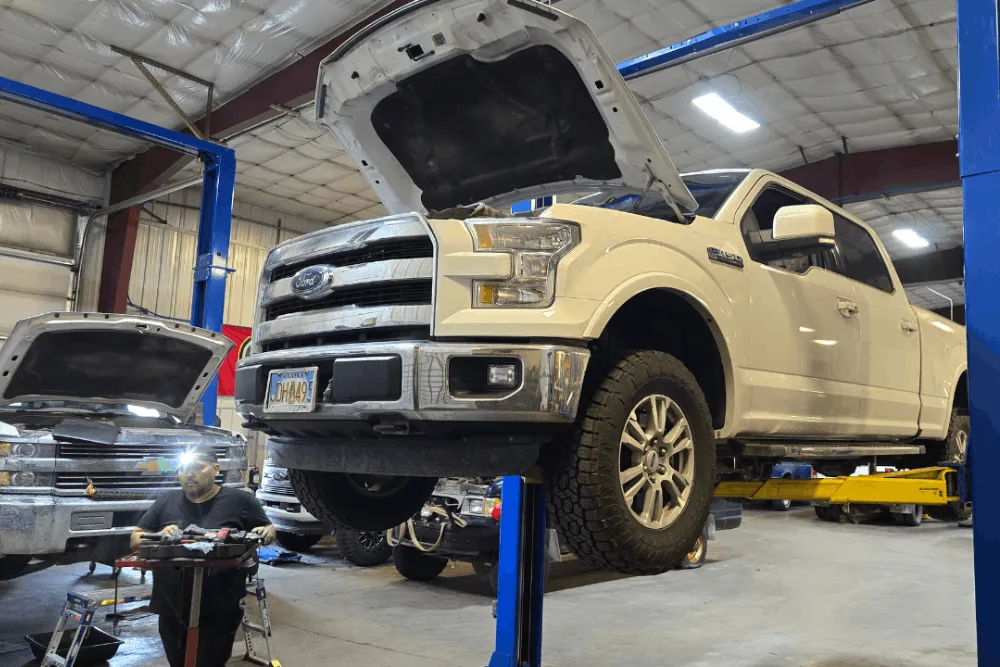 Towing Heavy in Alaska? Prevent Diesel Transmission & Cooling Failures - 1023 Diesel and Fleet - White pickup truck on a blue hydraulic lift in a large garage, hood open, with a mechanic working nearby. Brightly lit with a busy workshop vibe.