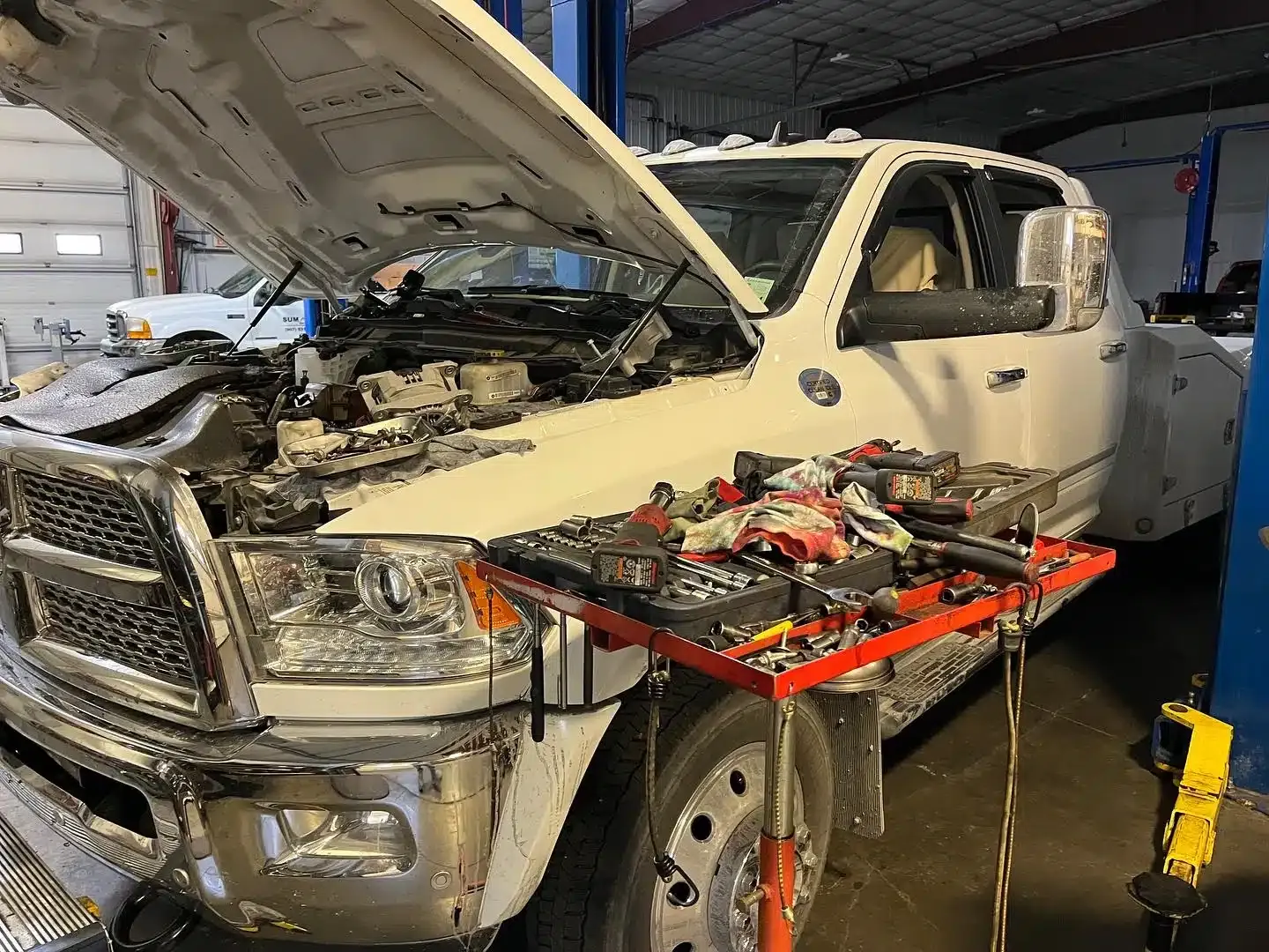 A white pickup truck with its hood open is parked inside a garage. Various tools and equipment are organized on a red tray attached to the front of the truck, indicating maintenance or repair work is underway.